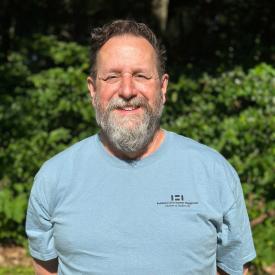 Bill Zito looks at the camera. He is standing outdoors with green bushes behind him and wearing a blue tee.