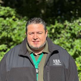 Carl Brokaw stands outside looking at the camera with green bushes behind him. He is wearing a green shirt and a black fleece jacket.