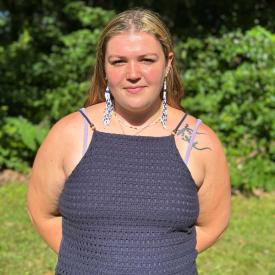 Juliette is looking at the camera, surrounded by a green outdoor backdrop. She is wearing a dark tank top. 