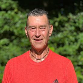 Kenny stares at the camera with a smile. He is standing outside, wearing a bright red tee.