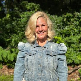 Lori is standing outdoors wearing a light denim jacket with puffy sleeves. She has short blonder hair and is smiling at the camera.