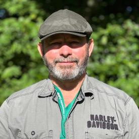 Mike is standing outdoors wearing a cap and a grey shirt.