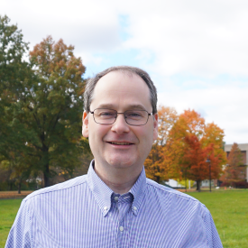 Michael Montgomery stands outside. He is wearing a blue shirt and glasses.