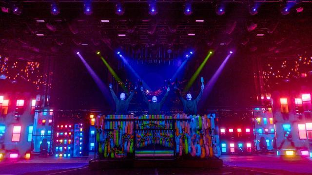  Three Blue Man Group performers stand behind a massive, colorful percussion instrument made of interconnected PVC pipes, raising their drumsticks in unison. The stage is illuminated by vibrant blue, purple, and green lighting, with spotlights shining down from above. The background features a dazzling digital cityscape with multicolored windows and abstract lighting effects, creating a futuristic and immersive atmosphere.