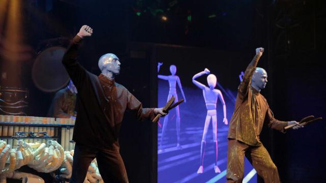  Two Blue Man Group performers, dressed in black and gold-splattered outfits with their faces painted blue, stand on stage raising their fists while holding wooden sticks. Behind them, a large screen displays animated humanoid figures mirroring their movements. Another performer in the background plays an elaborate percussion instrument made of interconnected PVC pipes. The stage lighting casts a dramatic glow, enhancing the theatrical and immersive atmosphere.