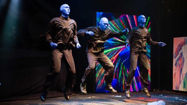 Three Blue Man Group performers, dressed in black with their faces and hands painted blue, leap into the air in unison during a stage performance. Their black clothing is splattered with paint. Behind them, a large screen displays a vibrant, swirling pattern of colors.