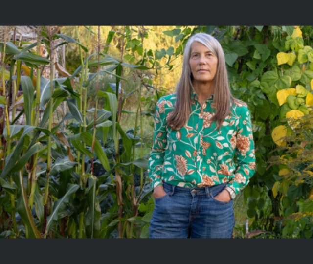 A person in a floral print shirt and jeans standing in front of a wall of plants.