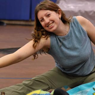 Alt text: A young woman with wavy brown hair and light skin smiles brightly while stretching on the floor. She is wearing a sleeveless blue top, olive green pants, and a silver necklace with small charms. 