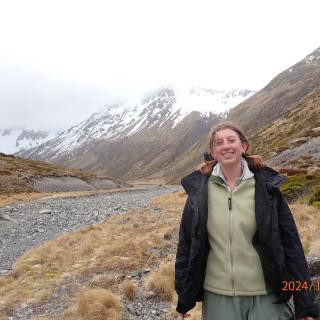 Alt text: A person with windblown hair smiles while standing in a rugged mountain landscape. They are wearing a light green fleece jacket and a black raincoat. Behind them, steep slopes covered in patches of grass and scattered rocks rise toward snow-capped peaks, partially obscured by clouds. A rocky riverbed winds through the valley. 