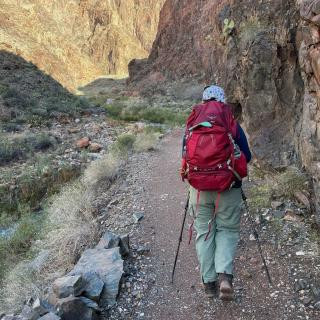 Alt text: A hiker wearing a floral-patterned hat, blue jacket, and green pants walks along a rocky trail in a canyon, carrying a large red backpack and using trekking poles. Towering reddish-brown canyon walls rise on either side, with patches of green vegetation clinging to the rocks. A small stream flows through the valley, and the sky above is bright blue.