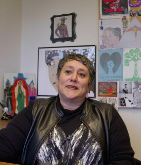 Professor of Critical Race, Gender, and Media Studies Susana Loza sits in her office. The background is full or colorful artwork.