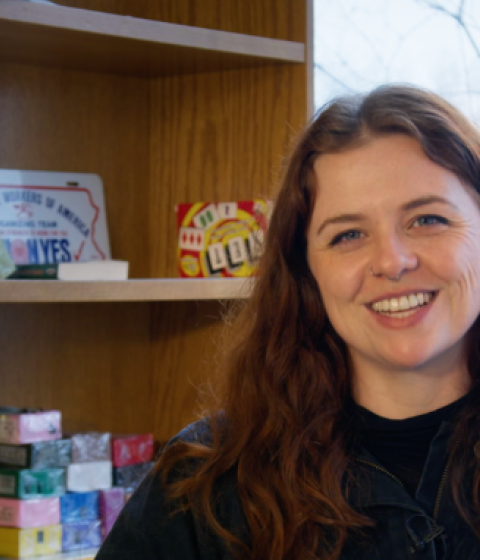 Sarah is sitting in a chair facing the camera. There is a wintery window behind her and a bookcase to her left.